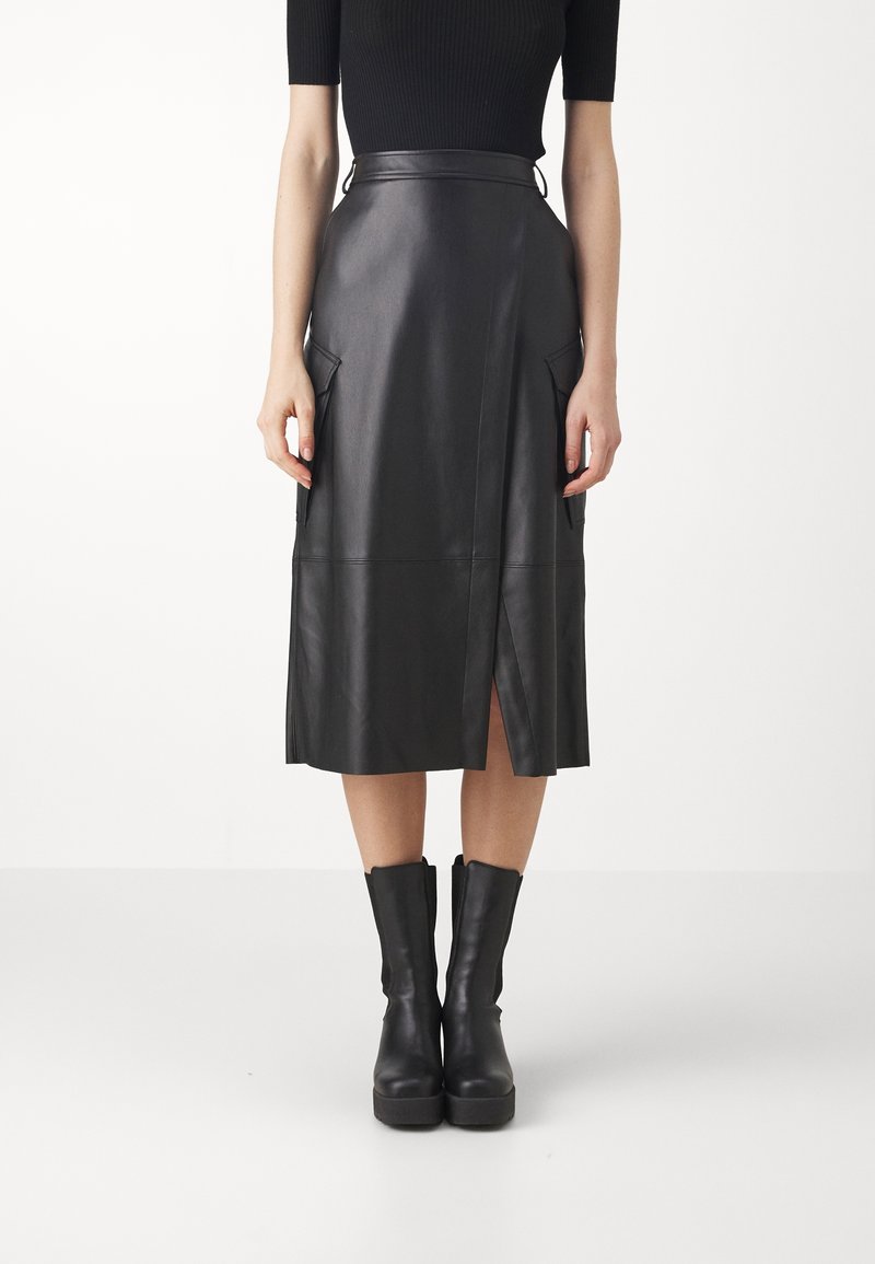 Femme portant une jupe midi en cuir noire avec une fente devant, des poches latérales à rabat, un top noir à manches courtes et des bottines noires sur fond blanc.