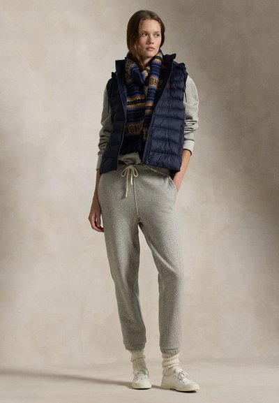 Jonge vrouw draagt grijze joggingbroek, grijze trui, marineblauw gewatteerd vest, patroon sjaal en witte sneakers met gebroken witte sokken.