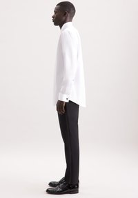 Camicia bianca a maniche lunghe con taglio dritto e colletto alto, abbinata a pantaloni neri su misura e scarpe nere lucide.
