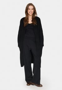 Lång svart cardigan i texturerat tyg, öppen front med fickor, kombinerad med åtsittande svart topp och vida svarta byxor. Bruna skor.