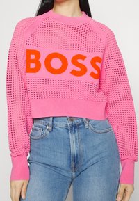 Růžový pletený svetr s otevřeným síťovaným vzorem, s výrazným oranžovým logem "BOSS" přes hrudník a žebrovanými manžetami na zápěstí.