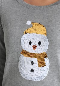 Pull gris avec un motif de bonhomme de neige en sequins. Le bonhomme de neige porte un chapeau doré et une écharpe marron, avec des boutons noirs et un nez rose.