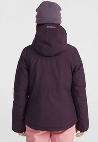 Tume lilla isoleeritud jakk kapuutsiga, millel on kaarjas äär ja väike logo seljal. Koos kergete roosade pükstega.