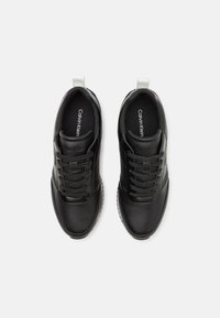 Zapatillas negras con parte superior de cuero suave, punta redonda, diseño con cordones, acentos blancos en el talón y suela de goma texturizada. Marca visible en el interior.