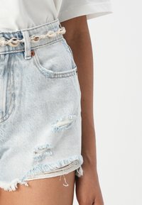Shorts en denim bleu clair, avec un ourlet effiloché, des accents usés, et une ceinture en coquillage blanche pour plus de détails.