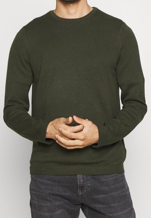 Mann trägt einen dunkelgrünen, langärmeligen Strickpullover mit gerippten Bündchen und Kragen, die Hände vor dem Körper verschränkt, kombiniert mit dunkelgrauen Jeans.