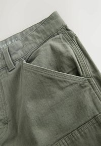 Green relaxed-fit pants featuring a smooth texture, a prominent side pocket, and visible stitching details on the waistband and pocket edge.