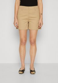 Beige Baumwollshorts mit mittelhohem Bund, Seitentaschen und geradem Beinabschluss, kombiniert mit dunklen Ballerinas mit goldener Akzentuierung.