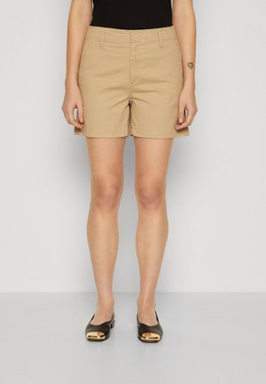 Shorts en coton beige à taille mi-haute, avec poches latérales et coupe droite, associés à des ballerines sombres avec un accent doré.