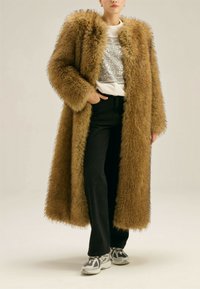 Osoba ubrana w długi brązowy płaszcz z sztucznego futra, białą koszulę w wzory, czarne spodnie i białe sneakersy, stojąca z jedną ręką w kieszeni.