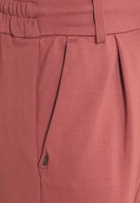 Närbild av lavendelfärgade byxor som visar elastisk midja, veckad front, bältesloop, sidoficka och liten svart märke.