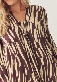Blouse marron et crème avec un motif de tourbillon abstrait. Présente un col en V et un détail noué à l'avant, fabriquée dans un tissu lisse.