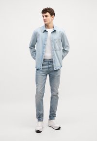 Camisa de denim azul claro con un bolsillo, llevada sobre una camiseta blanca, combinada con jeans azul claro y zapatillas blancas. Textura suave, corte casual.