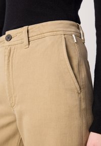 Khaki bomullbukser med frontlommer, metallknapplukking og sømdetaljering. Jevn tekstur med slim-fit design.