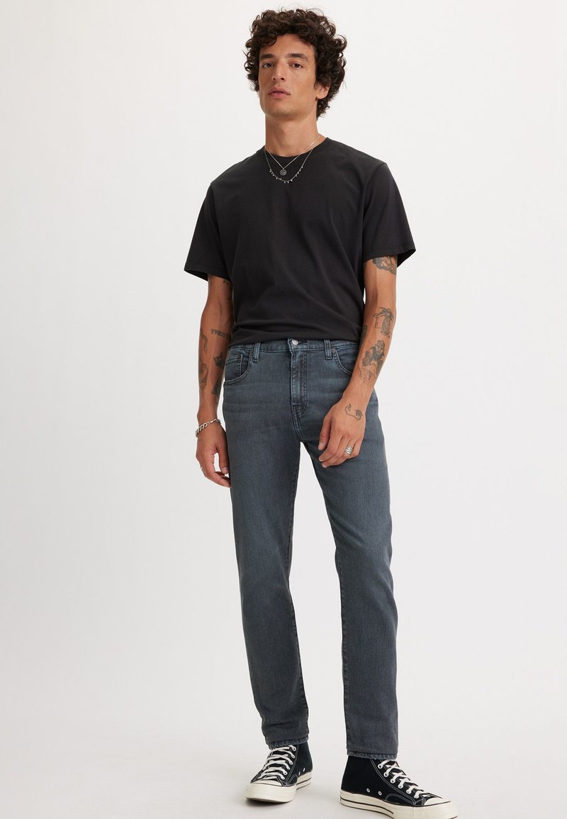 Schwarzes Kurzarm-T-Shirt kombiniert mit grauen Slim-Fit-Jeans. Auffällige Tätowierungen auf den Armen und schwarze Sneakers mit weißen Akzenten.