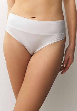 Femme portant un sous-vêtement blanc uni sans coutures à taille haute, debout avec le bras détendu le long du corps sur un fond neutre.