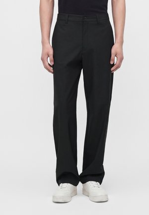 Homme portant un pantalon noir ample et des baskets blanches, debout devant un fond clair uni.