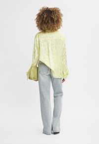 Camicia di raso verde lime con vestibilità ampia, maniche lunghe e orlo asimmetrico. Abbinata a jeans a gamba dritta blu chiaro e una piccola pochette.