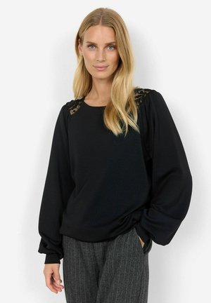 Pull noir avec des manches longues et volumineuses, orné de dentelle aux épaules. Tissu doux avec une coupe décontractée. Porté avec un pantalon gris à rayures.