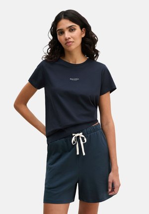Frau mit dunklem, welligem Haar, die ein marineblaues Marc O'Polo T-Shirt und passende Shorts mit weißem Kordelzug trägt und vor einem weißen Hintergrund steht.