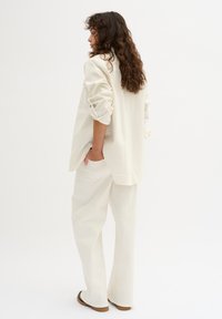 Camicia oversized color crema chiaro con maniche arrotolate e tasche frontali, abbinata a pantaloni larghi coordinati e scarpe piatte marroni su uno sfondo neutro.