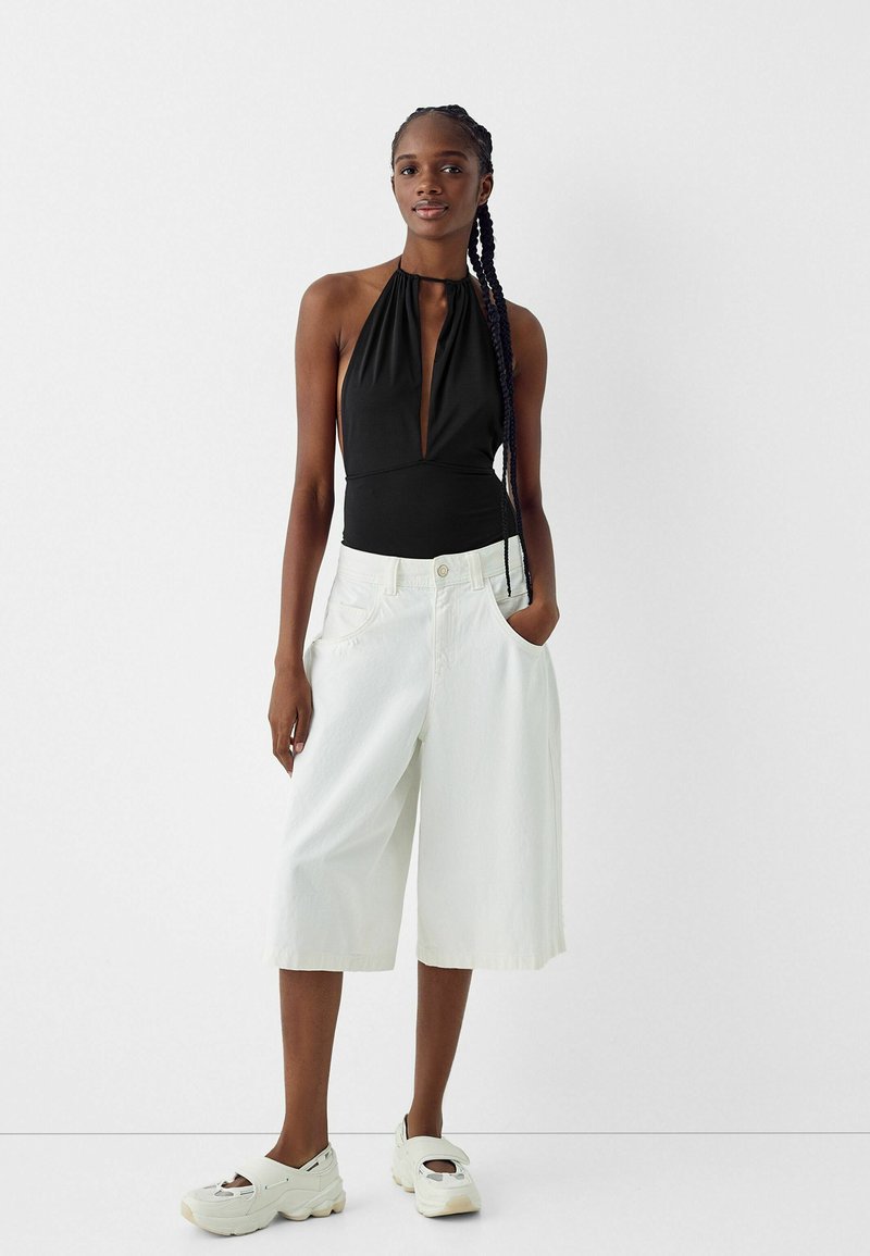 Schwarzer Halter-Bodysuit mit tiefem Ausschnitt, kombiniert mit weißen, weiten Shorts. Das Modell trägt weiße Slipper mit Ausschnitten.
