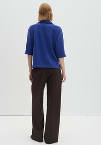 Personne aux cheveux mi-longs portant un haut bleu à manches courtes et un pantalon ample marron, debout face à un fond clair uni.