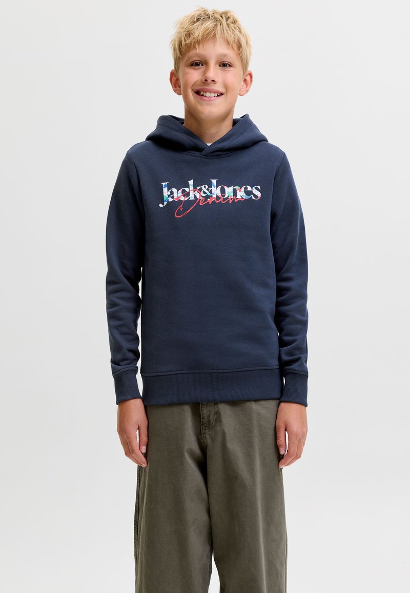 Garçon blond souriant portant un sweat à capuche bleu marine avec le texte "Jack & Jones" et un pantalon vert olive, debout contre un fond clair uni.