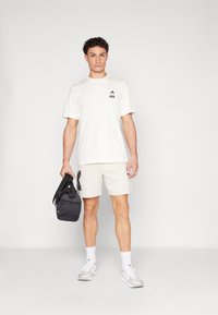 Κρέμ μπλουζάκι με μαύρο λογότυπο, συνδυασμένο με ανοιχτό μπεζ σορτσάκι με γραφικό. Το μοντέλο κρατάει μια μαύρη τσάντα, φορώντας άσπρες κάλτσες και γκρι παπούτσια.