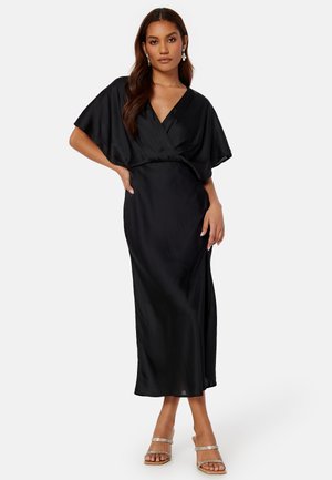 Abito avvolgente in satin nero con maniche a sbuffo corte, scollatura a V profonda, vita aderente e gonna fluida. Accentuato con sandali argento con cinturini.