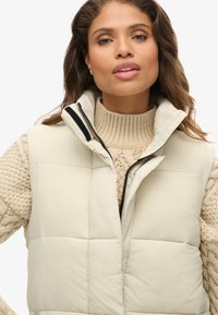 Gilet imbottito color crema con colletto alto, dotato di una cerniera frontale e dettagli neri, indossato sopra un maglione beige a maglia grossa.