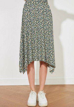 Kobieta w kwiatowej spódnicy midi z nierównym dołem i białych trampkach, stojąca na drewnianej podłodze na tle białej ściany.