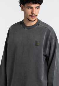 Sudadera gris con un cuello redondo, que presenta un pequeño parche de logo rectangular negro en el pecho. Textura suave y ajuste relajado.