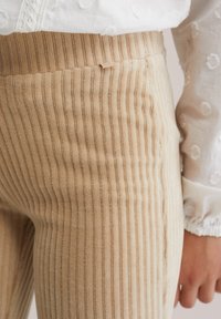 Bež hlače iz kordurove tkanine z navpičnimi rebrastimi progami, mehko teksturo in prilegajočo se zasnovo. Vključujejo majhno znamkasto etiketo blizu pasu.