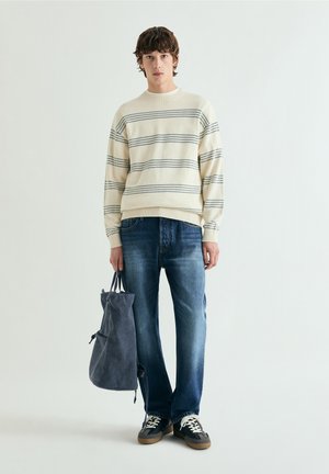 Giovane uomo con maglione crema a righe, jeans blu, sneakers nere, che tiene una borsa tote grigio scuro, in piedi davanti a uno sfondo chiaro e semplice.