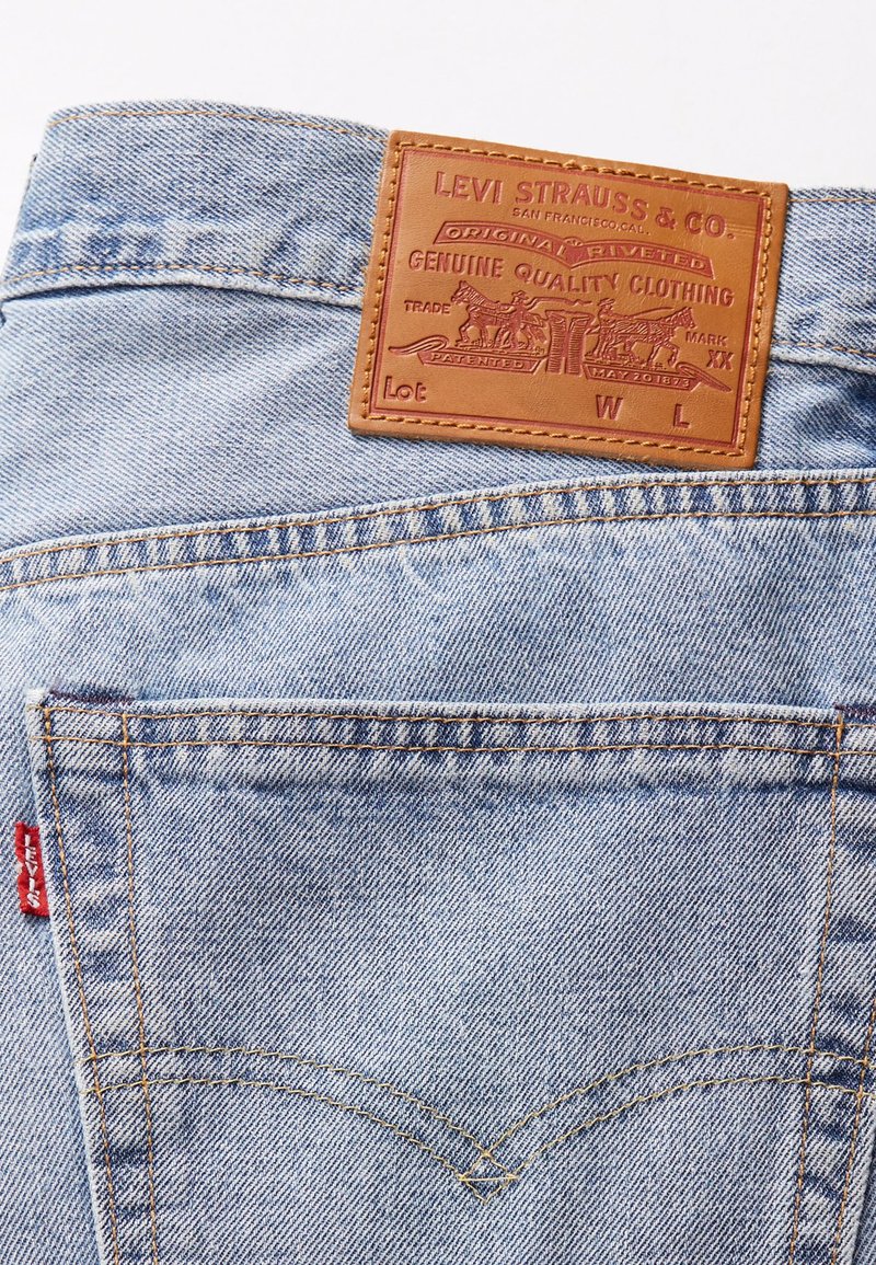Jeans de mezclilla azul claro con un parche de cuero marrón que presenta texto y diseños en relieve. Detalle de una etiqueta roja en el bolsillo lateral izquierdo.
