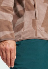 Beige fleece jack die een geometrisch patroon in lichtere tinten heeft, een zachte textuur en een contrasterende zoom. Hand rustend op teal broek.