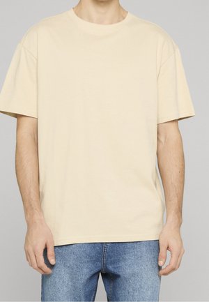 Mann trägt ein einfarbiges beiges T-Shirt mit kurzen Ärmeln und blaue Jeans, steht vor grauem Hintergrund, Oberkörper sichtbar ohne Kopf.