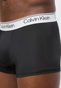 Κοντινό πλάνο ενός ατόμου που φοράει μαύρα ανδρικά εσώρουχα μποξεράκια με λευκή ελαστική μέση γερμένη με την επωνυμία «Calvin Klein».