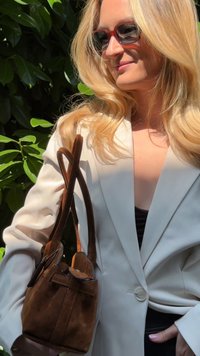 Bolso de ante marrón con asas de cuero; forma suave y estructurada; combinado con un blazer crema. Follaje verde en el fondo.