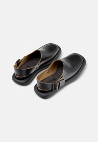 Scarpe slip-on nere con design a punta arrotondata, caratterizzate da una soletta a contrasto marrone e una cinturaregolabile nera. Texture liscia.
