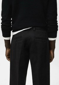 Schwarzer Strickpullover und schwarze Hose mit Gesäßtaschen und Knopfdetails. Der Pullover hat eine strukturierte Rippenoptik und darunter ein weißes, geschichtetes Hemd.