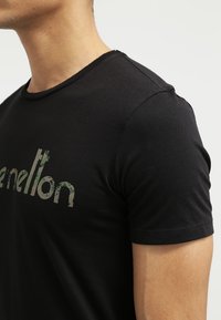 Černé tričko s krátkým rukávem a kulatým výstřihem, s mírně přiléhavým střihem, na přední straně s texturovaným, vybledlým zeleným logem.