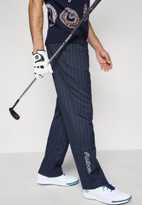 Tengerészkék csíkos golfnadrág laza szabással, fehér hímzett márkajelzéssel a szegélyen. Fehér golfcipővel és kesztyűvel viselve.