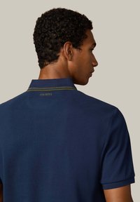 Granatowa koszulka polo z teksturowanej tkaniny, z krótkimi rękawami oraz kontrastowym akcentem w zielono-złotym kolorze na kołnierzyku, z nazwą marki "HACKETT" na plecach.