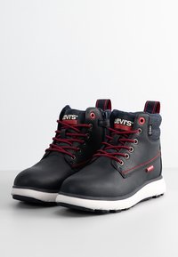 Bottines noires Levi's avec lacets rouges, semelles blanches, coutures rouges et col en tissu denim sur fond blanc uni.