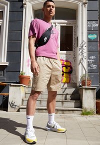 En vuxen man som bär en rosa bomullströja, beige shorts, vita Nike-strumpor och grå-gula Nike-skor. Han bär en svart midjeväska.