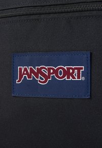 Juodas audinys su mėlyna etikete, ant kurios išsiuvinėtas "JANSPORT" logotipas bordo spalvos raidėmis. Užtrauktukas matomas virš etiketes.