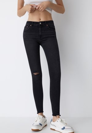 Vaqueros skinny negros con detalles desgastados en la rodilla derecha, dobladillo sin terminar en los tobillos, combinados con zapatillas blancas con acentos azules.