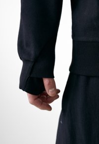 Fekete pulóver bordázott mandzsettákkal, amelyen egy kéz egy része látható, ahogy a szövetet markolja. Az anyag lágy, sima tapintású, és minimális varrással rendelkezik.